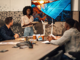 Portrait of multicultural ecology engineer standing with solar panels model in hands at boardroom and having seminar about clean and renewable energy development in front of diverse team of experts.