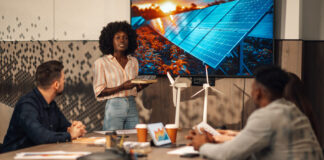 Portrait of multicultural ecology engineer standing with solar panels model in hands at boardroom and having seminar about clean and renewable energy development in front of diverse team of experts.