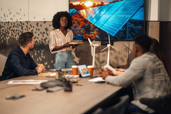 iStock-2170844691 (1) Portrait of multicultural ecology engineer standing with solar panels model in hands at boardroom and having seminar about clean and renewable energy development in front of diverse team of experts.