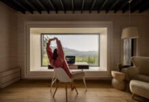 Woman stretching while works on laptop by the table in front of panoramic window with great view on mountains. Remote work and escaping to nature concept