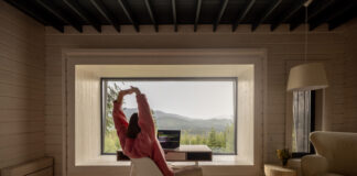 Woman stretching while works on laptop by the table in front of panoramic window with great view on mountains. Remote work and escaping to nature concept