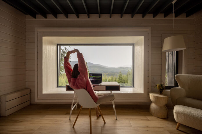 Woman works on laptop remotely in house on nature Woman stretching while works on laptop by the table in front of panoramic window with great view on mountains. Remote work and escaping to nature concept