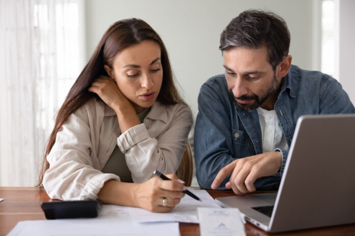 Wife and husband review financial documents seated at table with laptop, check receipts, discussing invoices, education savings or new rates.