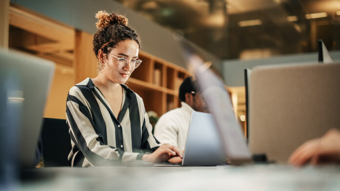 Diverse Group of Creative Colleagues Working on Laptops in Modern Office. Hispanic Female Graphics Designer Smiling and Typing. Colleagues Working on Projects in Background. Diverse Group of Creative Colleagues Working on Laptops in Modern Office.