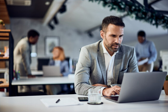 A focused businessman is working over the laptop, with people in the background. Photo of a focused businessman, dressed in a suit, working over the laptop, at the office. RTO Mirage concept