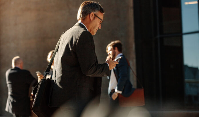 Man using mobile phone while walking to office Business people in hurry to reach office using mobile phone while walking on city street. People commuting to office in the morning carrying office bags busy with their mobile phones.