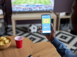 Close up of male hand holding smartphone with online sports bets on screen while watching football match at home,