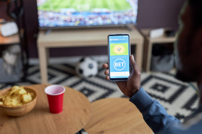 Close up of male hand holding smartphone with online sports bets on screen Close up of male hand holding smartphone with online sports bets on screen while watching football match at home,