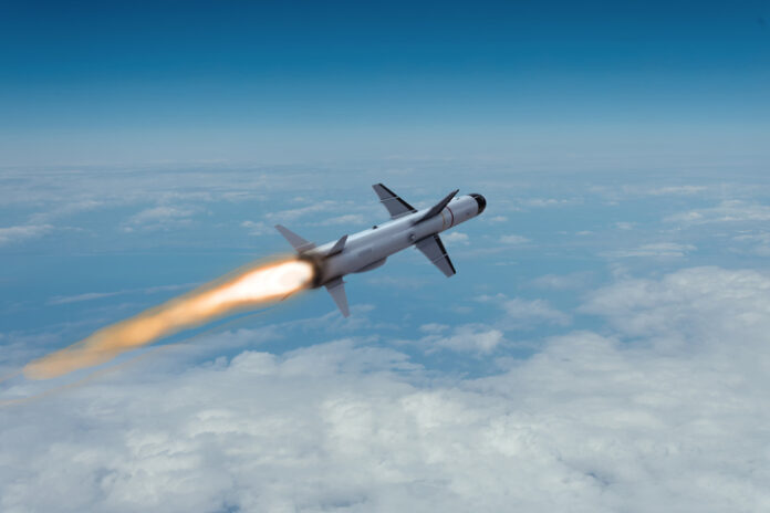 A modern anti-ship cruise missile flies at high altitude through the clouds, leaving a bright fiery trail in the blue sky. LCM cruise missile